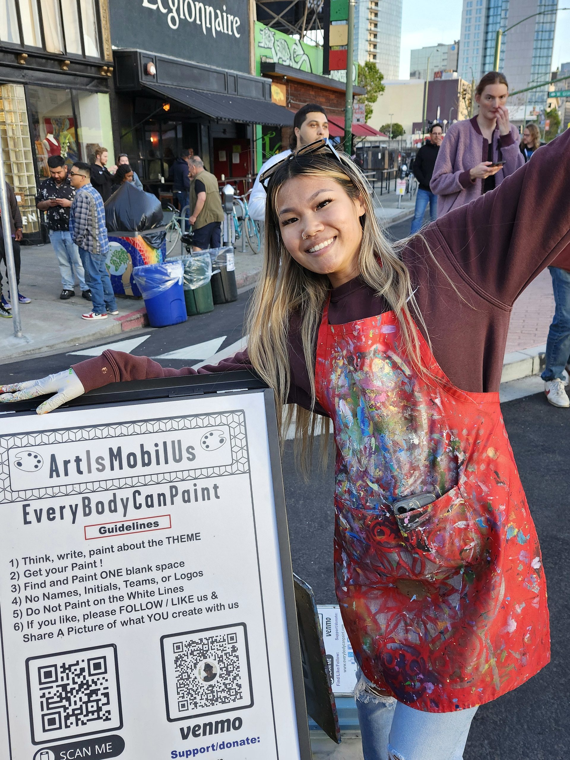 An Asian American woman with a painting apron stands next to a sign