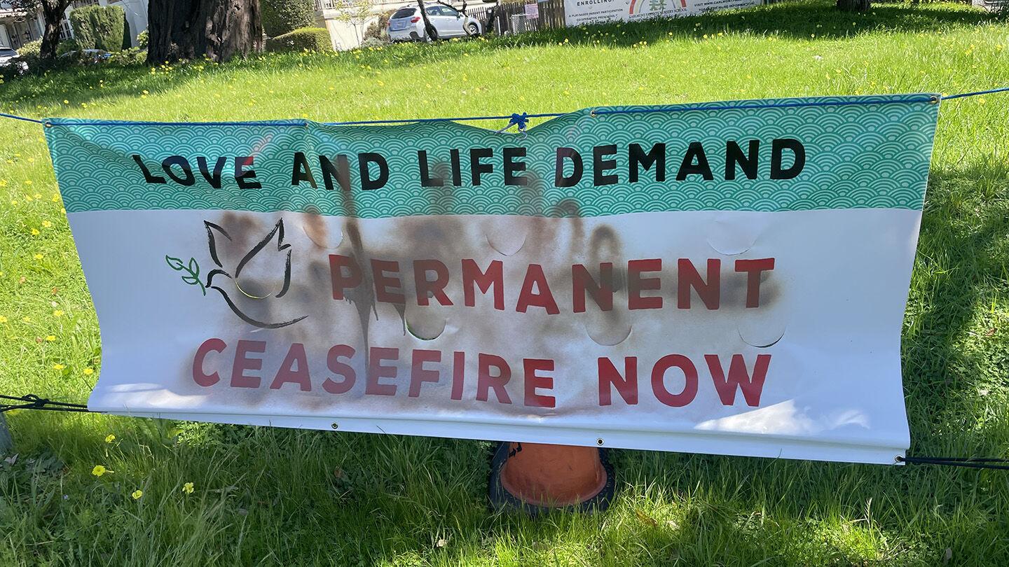 image of a white and teal banner that says "love demands ceasefire now" with brown spray paint over it