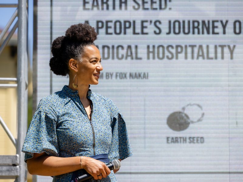 A Black woman stands on an outdoor stage with a microphone.