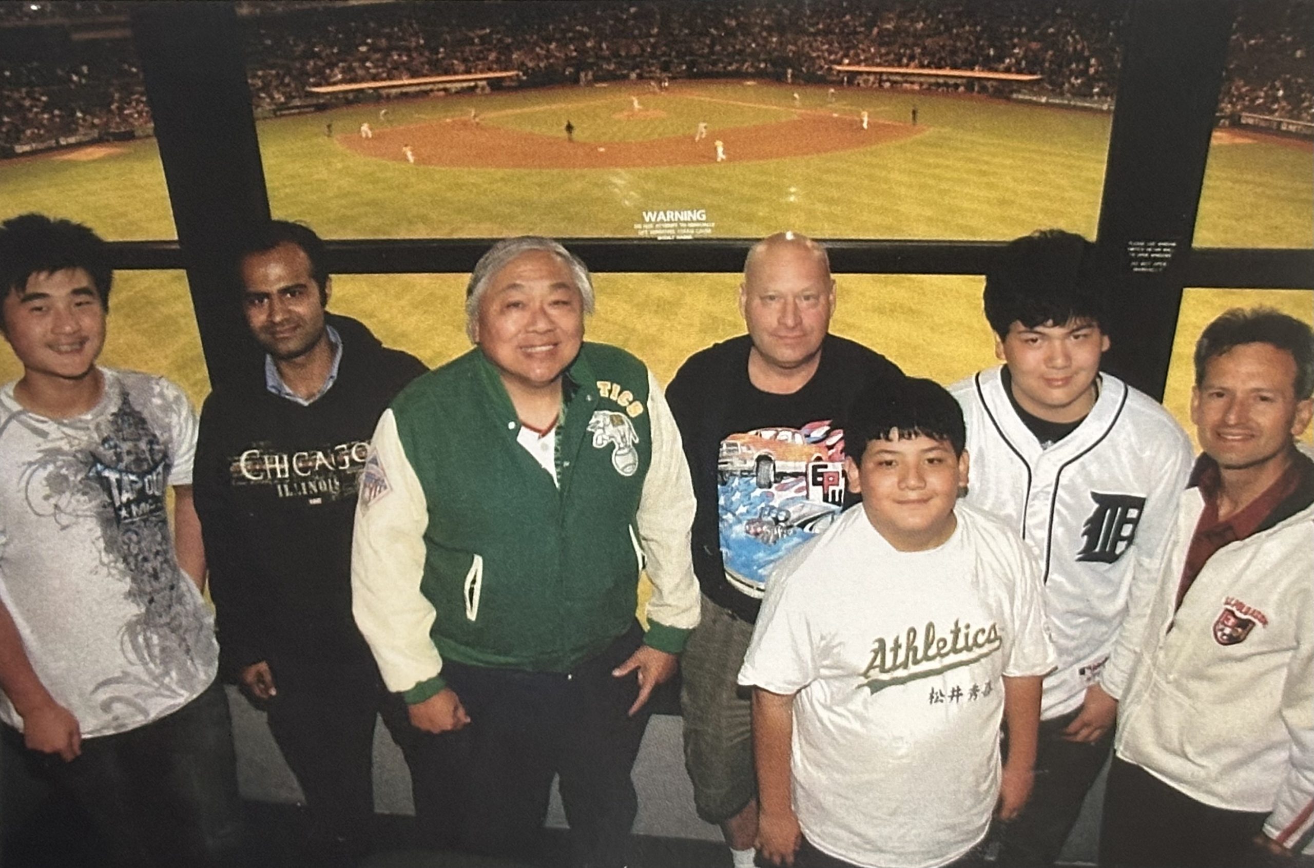Man stands with family and friends at baseball game. Baseball diamond in background.