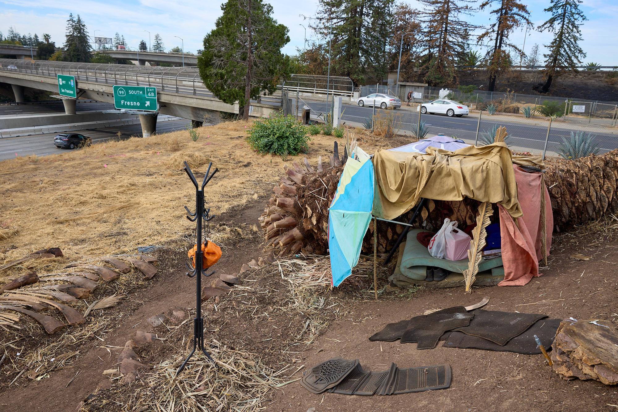 A small camp made form bed sheets and umbrellas sits on a hillside overlooking a highway with cars zooming by.