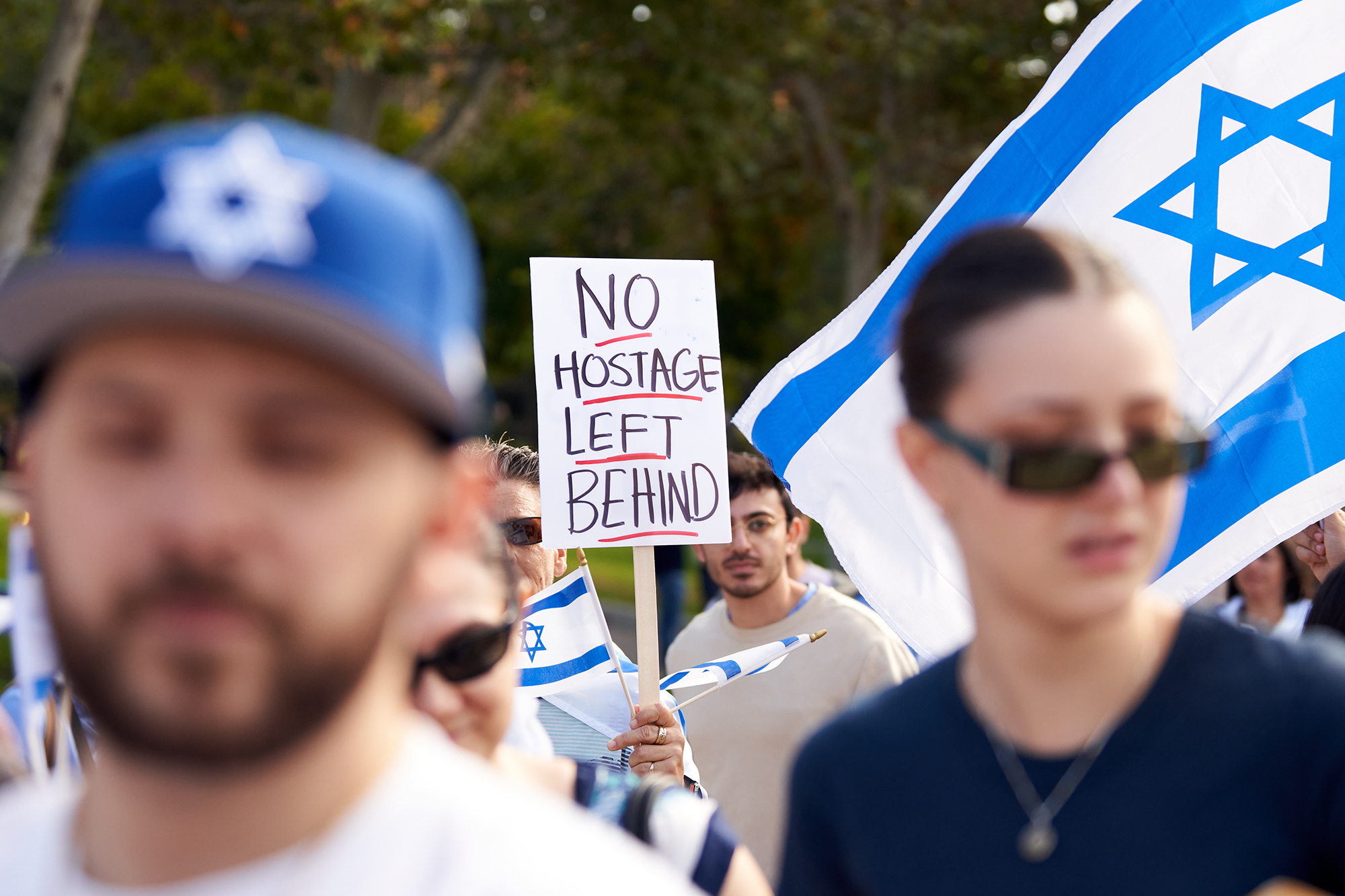 A person holds a sign reading "No Hostage Left Behind" during a rally, with Israeli flags visible in the crowd. The sign is in focus, while people and flags in the foreground are blurred. The event appears to be outdoors, with trees in the background.