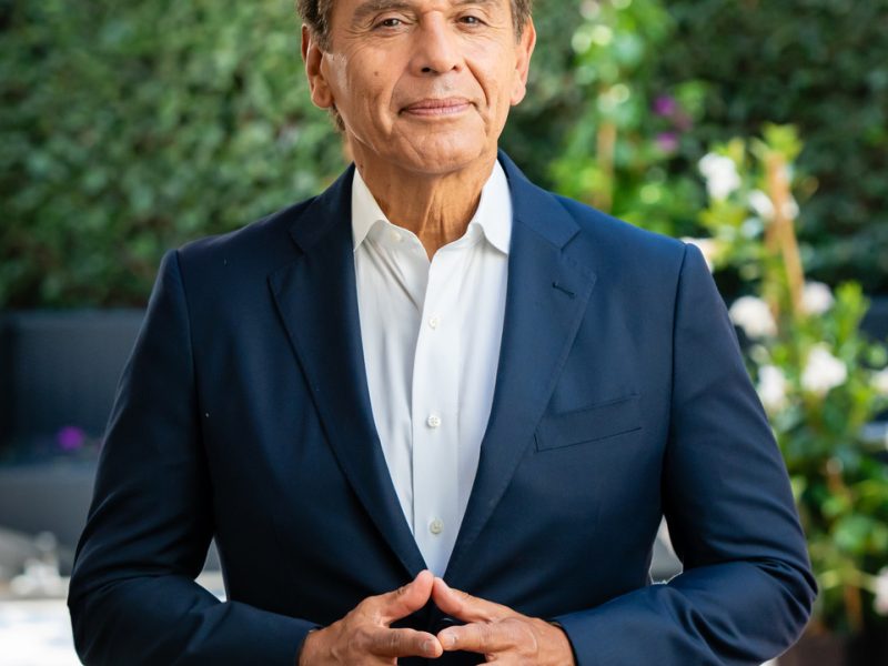 light brown latino mam wearing navy blue suit with open collar suit with slight smile and hands lightly held together