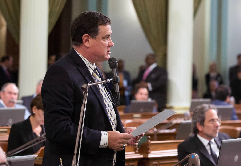A person stands at a microphone reading from a sheet of paper in a legislative chamber, with other people seated at desks in the background.