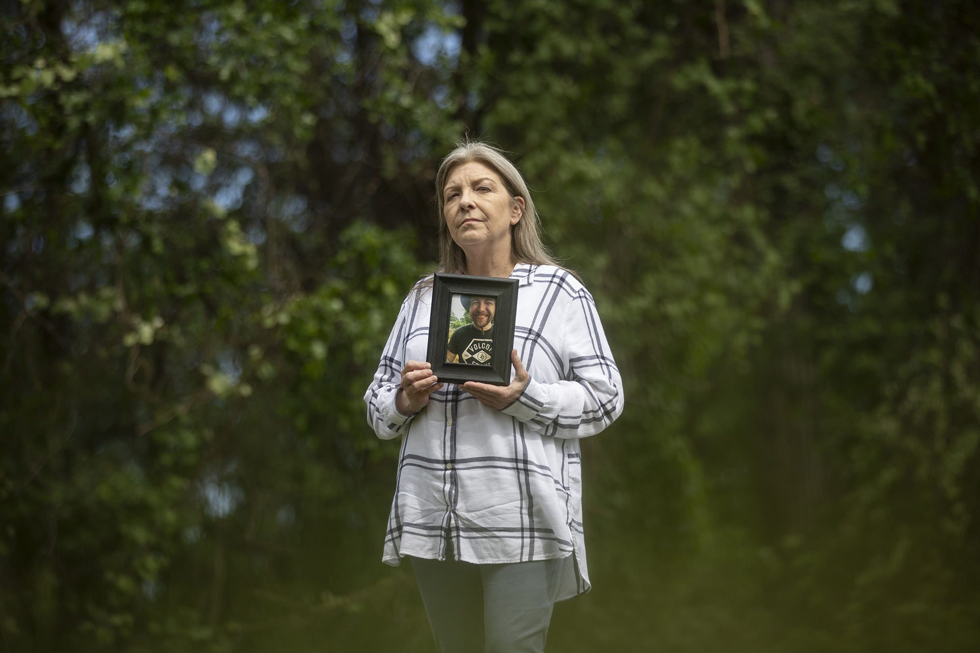 A person with grey hair wearing a white flannel shirt holds up a wooden framed photo of a person to their chest with both hands, In the background is a wall of green foliage from a row of trees behind the subject with specks of blue sky peaking though breaks in the branches.