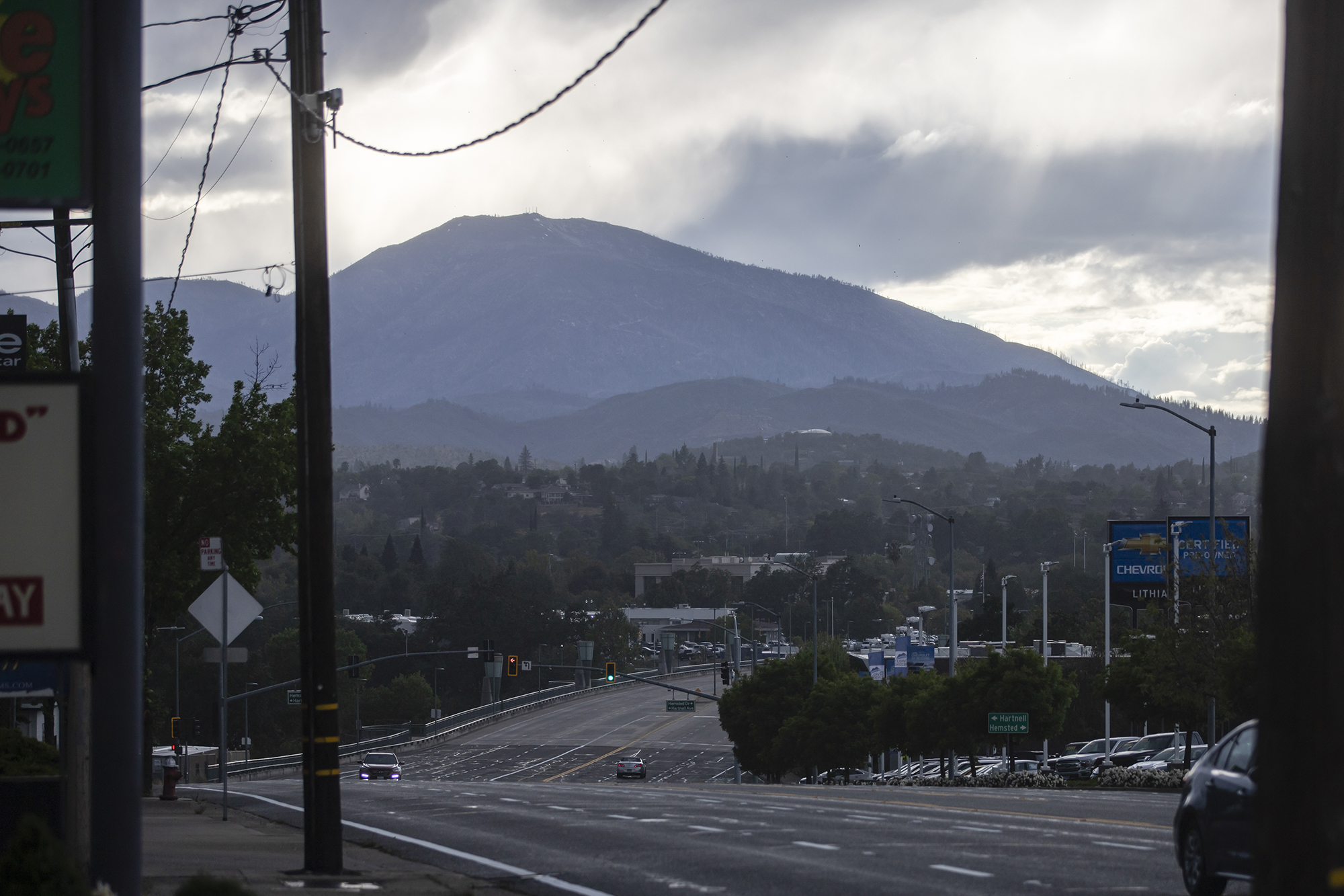 A view of a road leading into downtown Redding, with cars driving and a mountain range in the far background, while clouds create an overcast, giving a dark and gloomy scene.