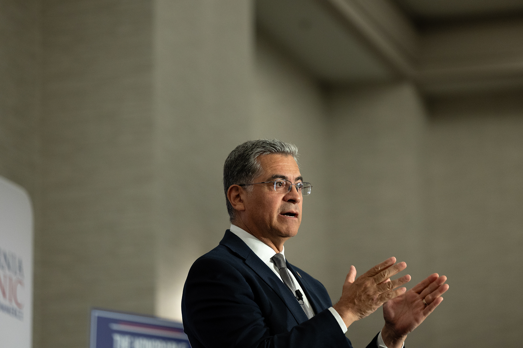 A side view of a person, with short gray hair and wearing a blue suit, gestures with both their hands as they stand on a small stage set against a beige wall inside a large room.