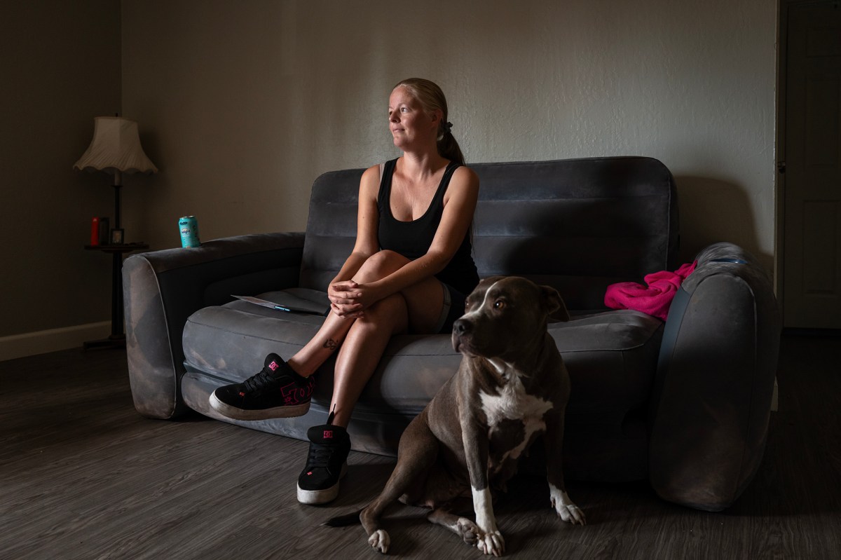 A woman sits on a couch in her apartment while her dog sits on the floor in front of her.