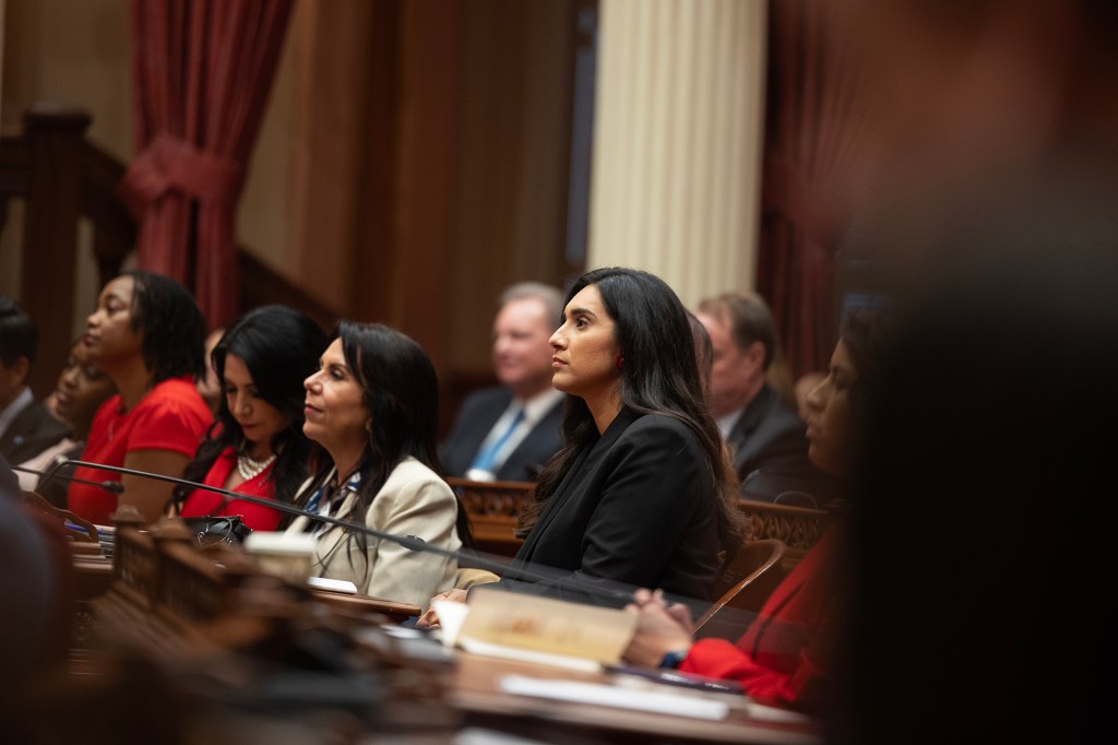 A person wearing a black blazer looks forward as they sit at a wooden desk, surrounded by other people.